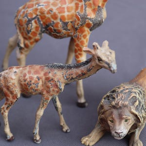 K&ouml;nnte beinhalten: Drei Vintage-Tierfiguren: eine Giraffe mit orangefarbenen und braunen Flecken, eine kleinere Giraffe und ein L&ouml;we. Der L&ouml;we befindet sich in einer geduckten Position mit detaillierter M&auml;hne und gr&uuml;nen Augen. Die Giraffen haben ein gealtertes Aussehen.
