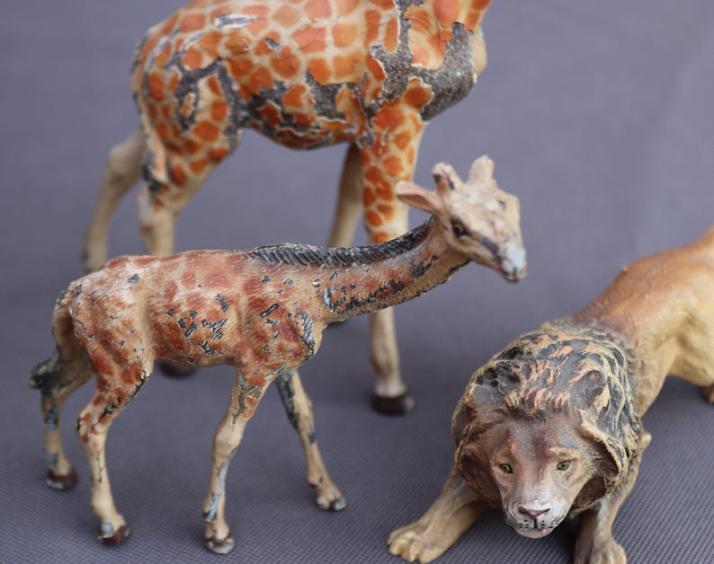 K&ouml;nnte beinhalten: Drei Vintage-Tierfiguren: eine Giraffe mit orangefarbenen und braunen Flecken, eine kleinere Giraffe und ein L&ouml;we. Der L&ouml;we befindet sich in einer geduckten Position mit detaillierter M&auml;hne und gr&uuml;nen Augen. Die Giraffen haben ein gealtertes Aussehen.