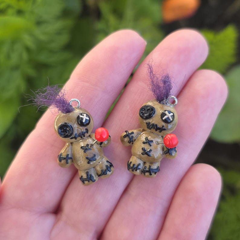 May include: Two handmade voodoo doll charms. Each doll is tan with black stitched details, button eyes, and a purple tuft of hair. A red pin is inserted into the doll's chest. The charms have a silver loop for hanging.