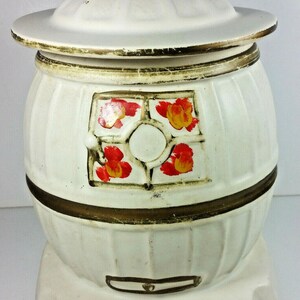 May include: White ceramic cookie jar shaped like a wood-burning stove with a gold trim and red and orange floral accents. The lid has a gold trim and a small knob on top.