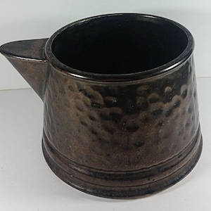 May include: A dark brown ceramic pitcher with a hammered texture. The pitcher has a wide mouth and a rounded body.