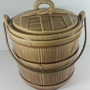 May include: A brown ceramic cookie jar shaped like a wooden bucket with a lid and a metal handle. The lid has a woven design and the bucket has a wood grain pattern.