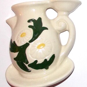 May include: A white ceramic wall vase with a green and white floral design. The vase has a handle and a spout, and it is shaped like a pitcher. It has a small plate on the bottom.