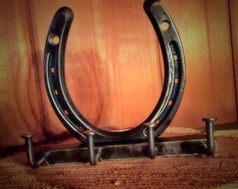 May include: A rustic horseshoe key holder made of dark metal. The horseshoe is mounted above a horizontal bar with three curved hooks. The key holder is displayed against a wooden background.