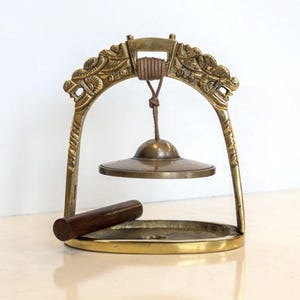 May include: A brass meditation chime with a decorative arch featuring ornate carvings. The chime includes a small, round cymbal suspended from a cord and a wooden striker resting in a circular base. The overall design is antique.