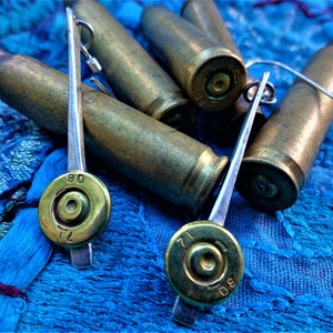 May include: A pair of silver earrings with bullet casings as the focal point. The casings are gold-toned and have the numbers "71" and "80" engraved on them. The earrings are hanging on a blue fabric background.