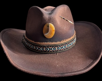 May include: A brown cowboy hat with a decorative band featuring a geometric pattern in black, turquoise, and gold. The hat has a brown stone and a feather accent. The hat is set against a black background.