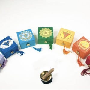 May include: A collection of seven square boxes in various colors, each adorned with a chakra symbol. The boxes have tassels and are arranged in a semi-circle. A small singing bowl with a wooden striker is in the center.