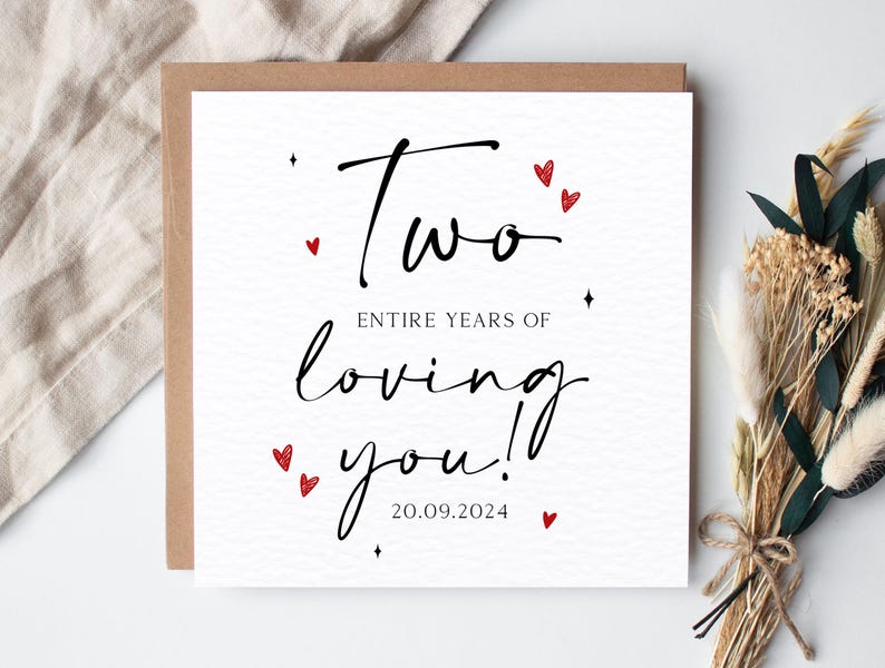 K&ouml;nnte beinhalten: Quadratische Gru&szlig;karte mit braunem Rand. Die wei&szlig;e Karte zeigt schwarzen Schreibschrifttext mit der Aufschrift &bdquo;Two entire years of loving you!&ldquo; mit roten Herzakzenten und dem Datum 20.09.2024. Trockenblumen im Hintergrund.