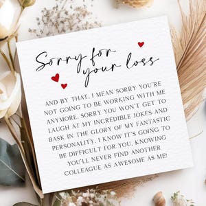May include: A white card with the text "Sorry for your loss" in black script, with three red heart accents. The card's message continues in a smaller, black sans-serif font. The card is surrounded by white flowers and dried foliage.