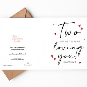 K&ouml;nnte beinhalten: Wei&szlig;e Gru&szlig;karte mit dem Text "Two entire years of loving you!" in schwarzer Schrift, verziert mit roten Herzen und dem Datum 20.09.2024. Die Karte ist innen leer und wird mit einem braunen Umschlag geliefert.