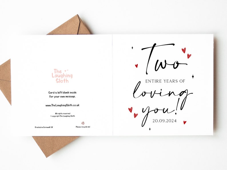 K&ouml;nnte beinhalten: Wei&szlig;e Gru&szlig;karte mit dem Text "Two entire years of loving you!" in schwarzer Schrift, verziert mit roten Herzen und dem Datum 20.09.2024. Die Karte ist innen leer und wird mit einem braunen Umschlag geliefert.