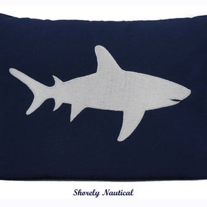 May include: Navy blue rectangular pillow featuring a white shark design. The shark is embroidered on the pillow, which has a solid color background. The text "Shorely Nautical" is at the bottom of the pillow.