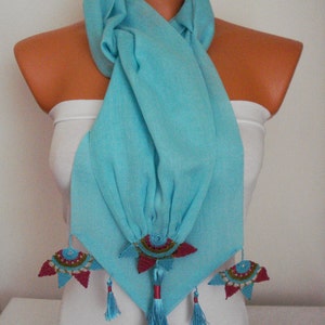 May include: A light blue scarf with decorative floral accents. The scarf is draped over a mannequin and features intricate, colorful embellishments. The embellishments include red, teal, and gold details, with matching tassels. The scarf is made of a soft, flowing fabric.