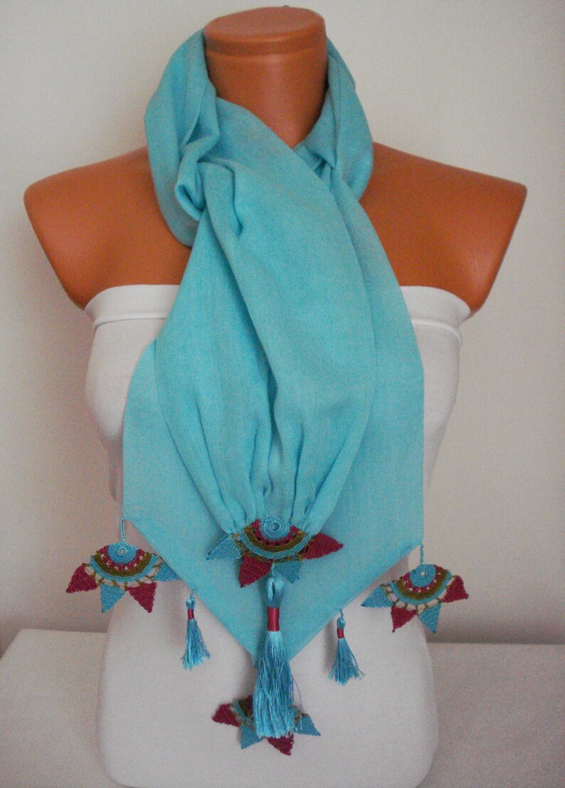 May include: A light blue scarf with decorative floral accents. The scarf is draped over a mannequin and features intricate, colorful embellishments. The embellishments include red, teal, and gold details, with matching tassels. The scarf is made of a soft, flowing fabric.