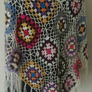 May include: A crocheted poncho featuring a colourful granny square pattern and white fringe. The poncho is made in a range of colours, including pink, purple, blue, green, yellow and orange.