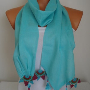 May include: A turquoise scarf with decorative embellishments. The scarf is draped over a mannequin, showcasing its flowing fabric. The ends feature colorful, embroidered accents and matching tassels, adding a touch of elegance to this accessory.