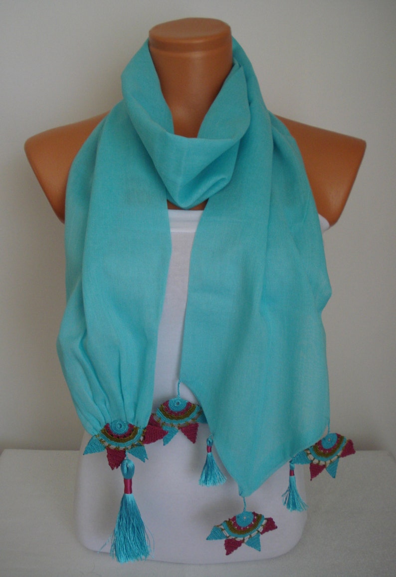 May include: A turquoise scarf with decorative embellishments. The scarf is draped over a mannequin, showcasing its flowing fabric. The ends feature colorful, embroidered accents and matching tassels, adding a touch of elegance to this accessory.
