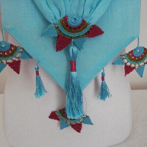 May include: A turquoise fabric with decorative crochet embellishments in red, teal, and gold. The design includes tassels and star-shaped crochet elements, creating a unique textile accent.
