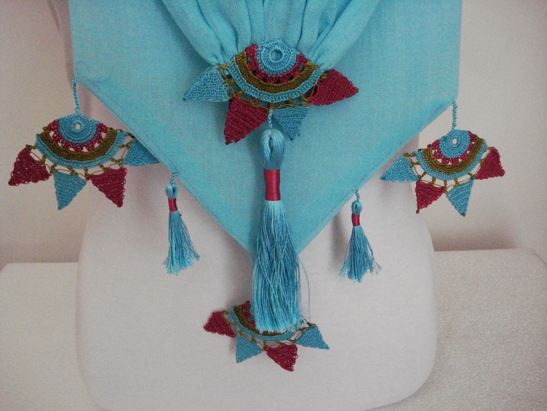 May include: A turquoise fabric with decorative crochet embellishments in red, teal, and gold. The design includes tassels and star-shaped crochet elements, creating a unique textile accent.