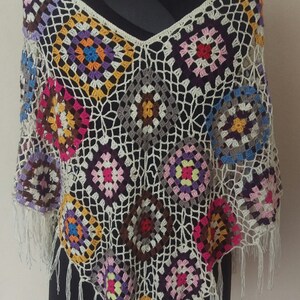 May include: A crocheted poncho with a white background and colourful geometric patterns. The poncho has fringe around the edges.