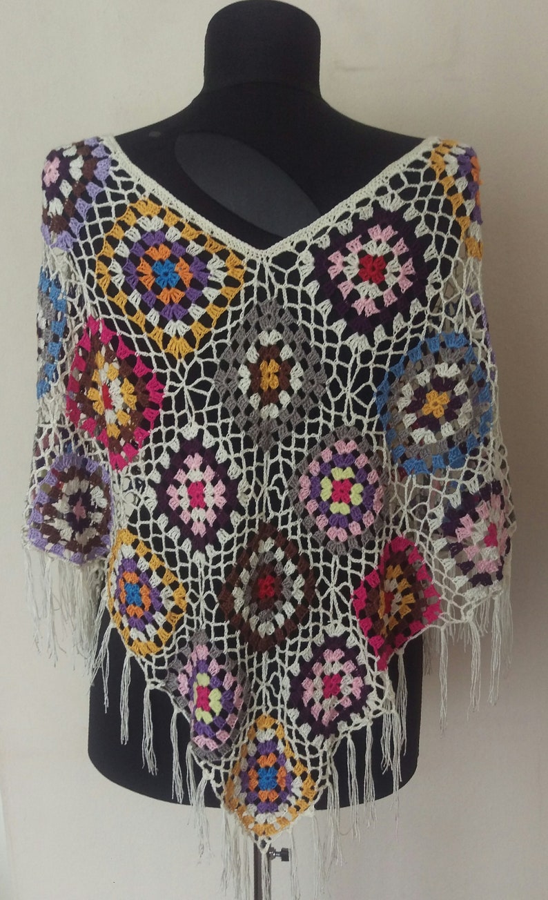 May include: A crocheted poncho with a white background and colourful geometric patterns. The poncho has fringe around the edges.