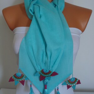May include: A turquoise scarf with decorative floral accents. The scarf is draped over a mannequin, showcasing its flowing fabric and intricate design. The floral accents are a combination of pink, teal, and gold, with matching tassels.