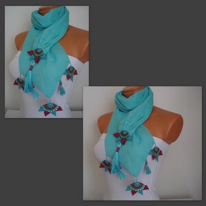 May include: A turquoise scarf with decorative embellishments. The scarf features star-shaped accents with red and teal details, and dangling tassels. The scarf is draped on a mannequin.