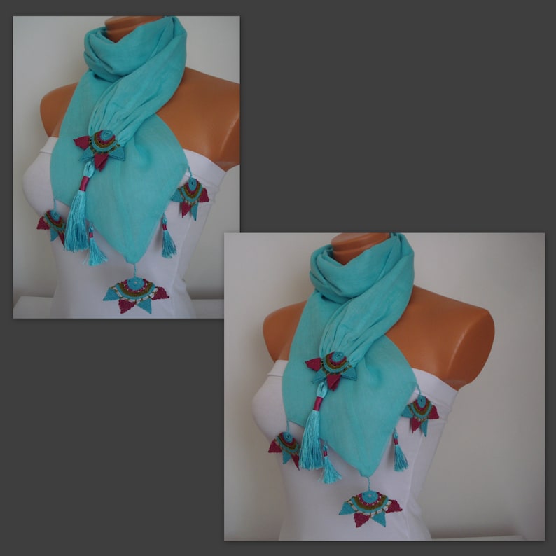 May include: A turquoise scarf with decorative embellishments. The scarf features star-shaped accents with red and teal details, and dangling tassels. The scarf is draped on a mannequin.