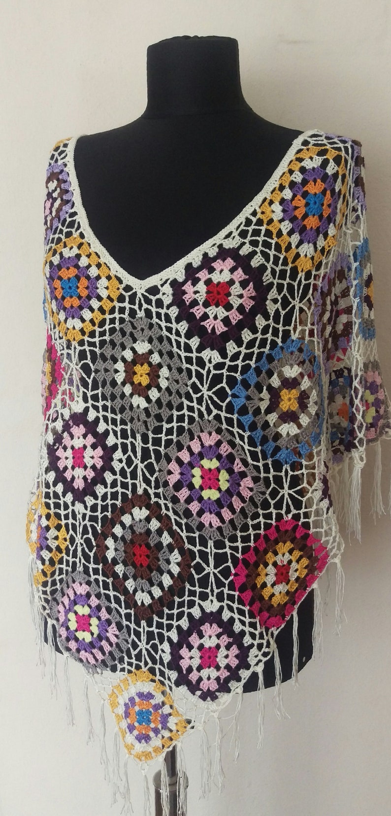 May include: A white crochet poncho with colourful granny squares. The poncho has a v-neck and fringe trim.
