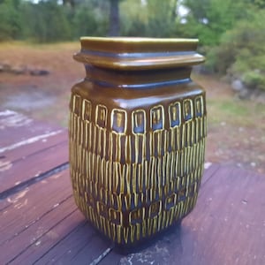 May include: A brown ceramic vase with a textured, geometric pattern. The vase has a rectangular shape with a slightly flared top and a series of horizontal ridges. The design features vertical lines and rectangular shapes.