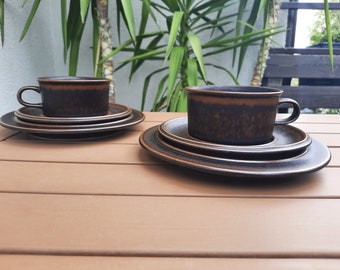 以下が含まれることがあります： 茶色い陶器のティーカップとソーサーの2セットが、茶色の木製のテーブルに配置されています。カップは濃い茶色の外観で、縁の近くに明るい茶色の帯があります。ソーサーは濃い茶色で、明るい茶色の縁があります。背景には緑の植物があります。