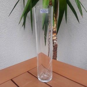 May include: A tall, clear glass vase with a decorative etched design. The vase has a cylindrical shape and is approximately 12 inches tall. The vase is sitting on a brown surface.