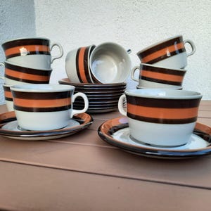 Puede incluir: Un juego de tazas y platillos de café vintage. Las tazas y los platillos son blancos con rayas horizontales marrones y naranjas. Las tazas tienen un asa y están apiladas. Los platillos son redondos y tienen un borde marrón.