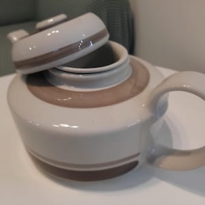 May include: A white ceramic teapot with a brown stripe design. The lid is off and the teapot is sitting on a white surface.