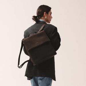 May include: A dark brown and black backpack with a flap closure. The backpack has adjustable straps and is worn over the shoulders. The person in the image is wearing a dark gray blazer, blue jeans, and black shoes.