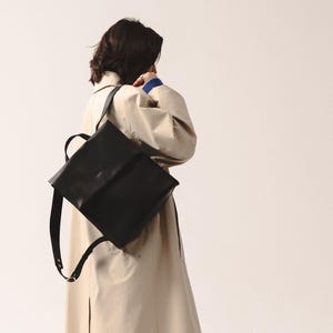 woman in a beige trench coat wearing a black Fi leather backpack with structured square shape and adjustable straps, styled for an elegant urban look