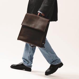 Woman in dark blazer and jeans holding a dark brown leather backpack with flap closure by Taksuba.