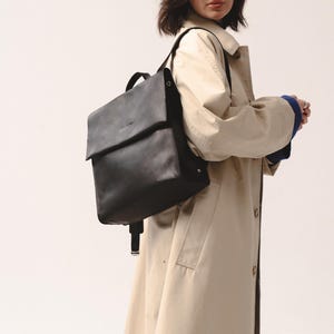 May include: A black leather backpack with a flap closure. The backpack has adjustable straps and a top handle. The person is wearing a tan trench coat with a blue sweater underneath.