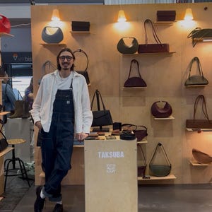 May include: A display of leather handbags in various shapes and colors, including olive green, burgundy, and black. The bags are arranged on wooden shelves with warm lighting. The brand name, TAKSUBA, is visible.
