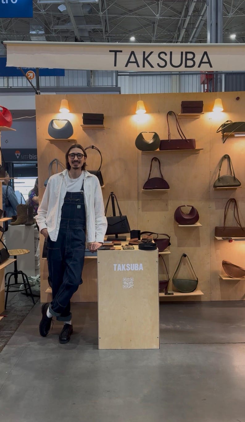 May include: A display of leather handbags in various shapes and colors, including olive green, burgundy, and black. The bags are arranged on wooden shelves with warm lighting. The brand name, TAKSUBA, is visible.