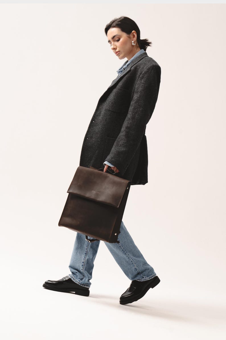 Woman in dark blazer and jeans holding a dark brown leather backpack with flap closure by Taksuba.