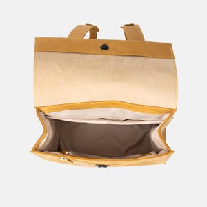 May include: Open view of a mustard yellow backpack with a flap closure and adjustable straps. The interior is a light beige color with a zippered pocket. The bag is made of a soft, textured material.