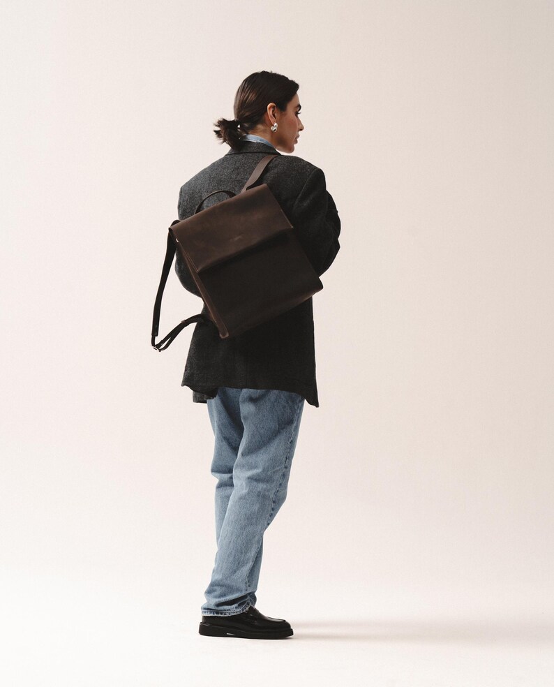 May include: A dark brown and black backpack with a flap closure. The backpack has adjustable straps and is worn over the shoulders. The person in the image is wearing a dark gray blazer, blue jeans, and black shoes.