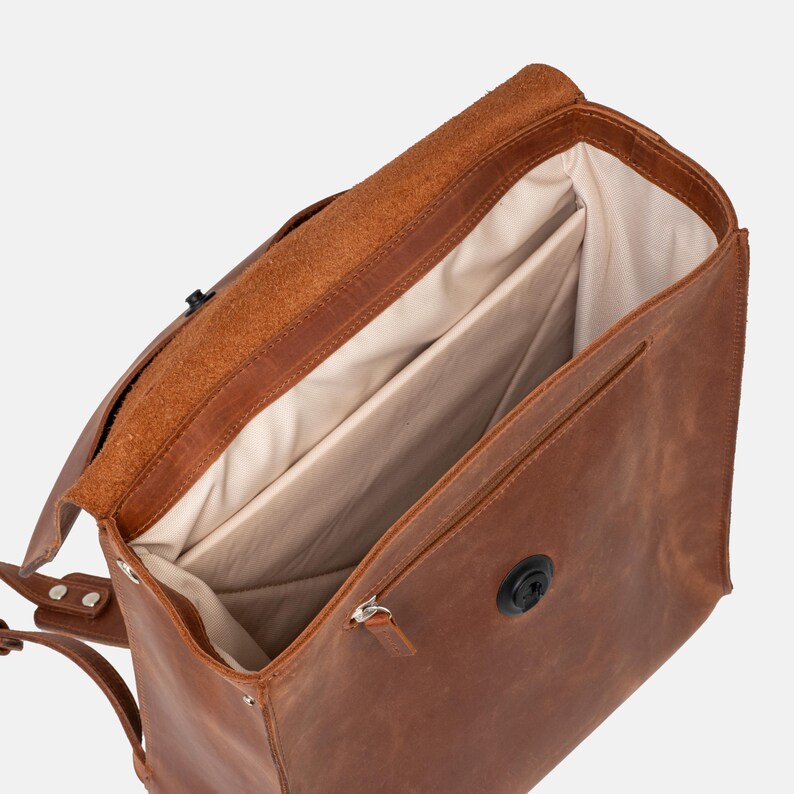 May include: A brown leather backpack with an open flap, revealing a light-colored interior with multiple compartments. The bag features a black clasp and zipper, and adjustable straps. The leather has a textured appearance.