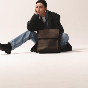 May include: A dark brown leather backpack with a flap closure. The bag has a rectangular shape and is sitting on a white surface. The backpack has adjustable straps and a top handle. The bag is a neutral color and can be used for everyday use.