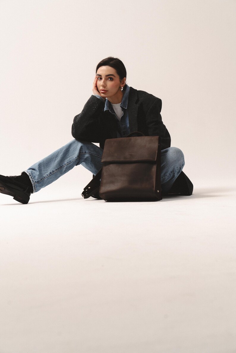 May include: A dark brown leather backpack with a flap closure. The bag has a rectangular shape and is sitting on a white surface. The backpack has adjustable straps and a top handle. The bag is a neutral color and can be used for everyday use.