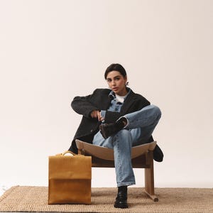 May include: A mustard-colored leather briefcase with a flap closure sits on a woven rug. The briefcase is rectangular with a handle on top. The image shows a person wearing a dark gray blazer, denim shirt, and jeans.