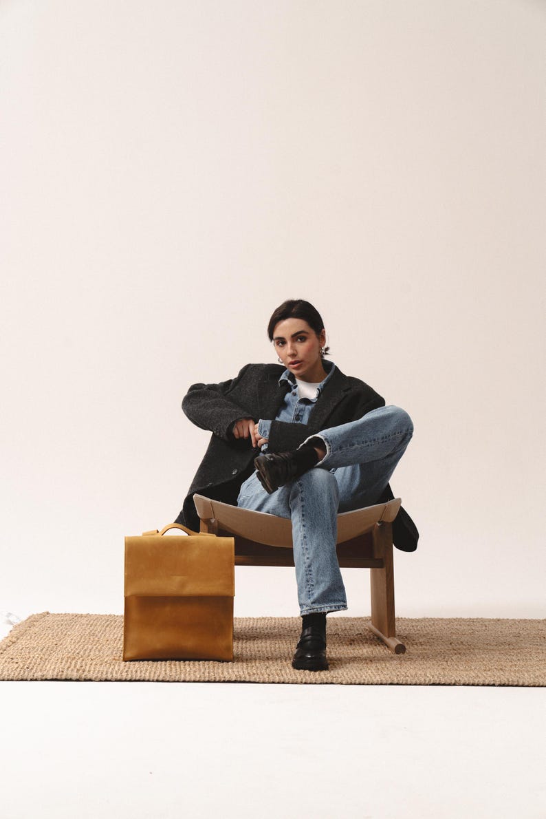 May include: A mustard-colored leather briefcase with a flap closure sits on a woven rug. The briefcase is rectangular with a handle on top. The image shows a person wearing a dark gray blazer, denim shirt, and jeans.