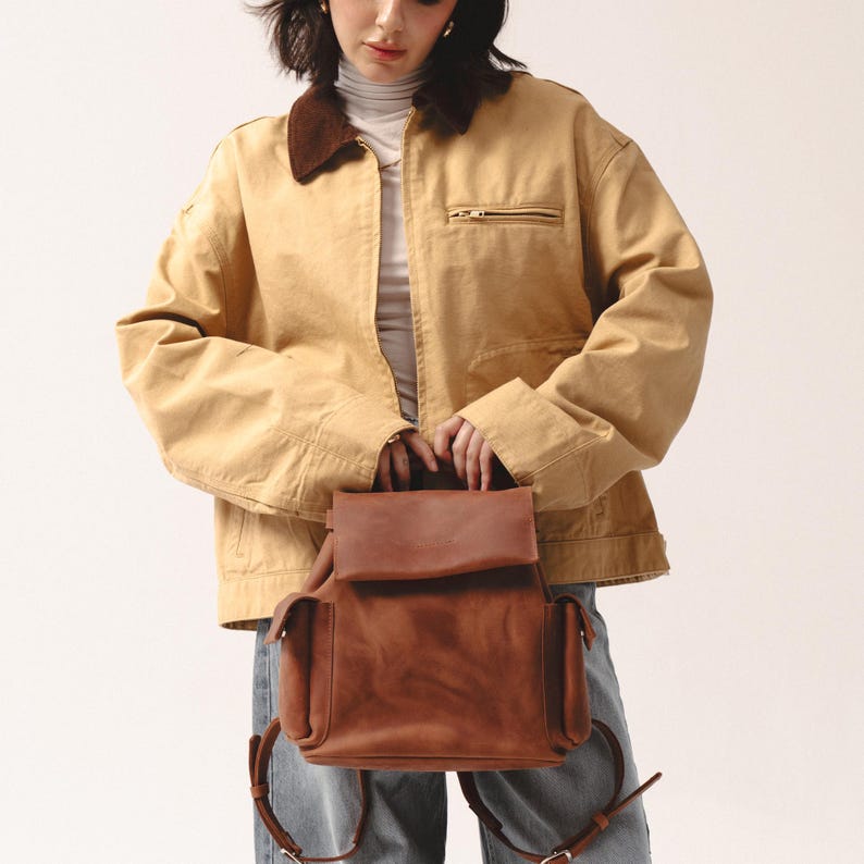 woman in a yellow jacket holding a small brown European leather backpack with flap closure, top handle, and visible side pockets, styled for everyday casual wear